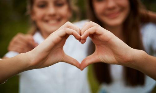 Love hand sign, happy sisters in garden with fun relationship, bonding and care with siblings. Heart hands, girls and nature, kindness and friendship for teenagers in park for quality time together